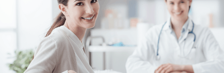 Paciente e médica em consultório. Ambas estão olhando para a foto e sorrindo. A paciente está de costas e virou para a imagem.